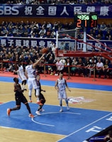 A basketball game takes place in a large indoor arena filled with spectators. Players from two teams, one in black uniforms and the other in white, compete for the ball near the hoop. The stadium is adorned with banners in Chinese, and a scoreboard displays a tied score of 21. The court features a distinctive blue and wood pattern.