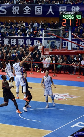 A basketball game takes place in a large indoor arena filled with spectators. Players from two teams, one in black uniforms and the other in white, compete for the ball near the hoop. The stadium is adorned with banners in Chinese, and a scoreboard displays a tied score of 21. The court features a distinctive blue and wood pattern.