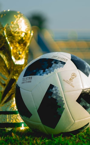 A gold trophy and a black-and-white soccer ball are placed on a green grass field. The background shows blurred stadium seating, suggesting a sports venue.