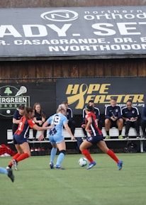 Players engage in a competitive soccer game on a field while others are seated on the bench watching attentively. The action is taking place in front of a wooden structure displaying multiple advertisements, including orthodontics, lacrosse, and field hockey.
