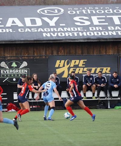 Players engage in a competitive soccer game on a field while others are seated on the bench watching attentively. The action is taking place in front of a wooden structure displaying multiple advertisements, including orthodontics, lacrosse, and field hockey.