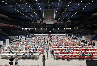 An indoor sports arena filled with rows of tables, each occupied by people engaged in competitive activities. The ceiling is high, with numerous lights illuminating the large space. The audience seating on either side is empty, and there are flags hanging from the upper level.