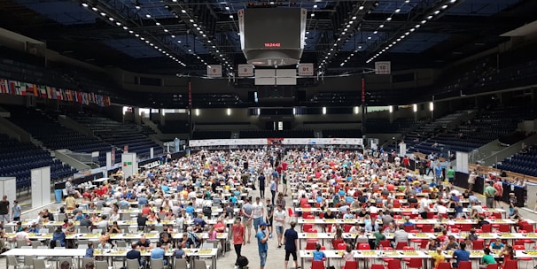 An indoor sports arena filled with rows of tables, each occupied by people engaged in competitive activities. The ceiling is high, with numerous lights illuminating the large space. The audience seating on either side is empty, and there are flags hanging from the upper level.