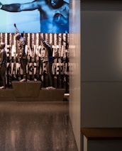 A dimly lit display features statues of athletes standing on a podium. They are positioned in front of a backdrop with the words 'LEVELING THE PLAYING FIELD' in bold letters. On the wall to the right, a softly lit quote is partially visible. The overall ambiance suggests a museum or exhibition setting.