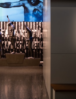 A dimly lit display features statues of athletes standing on a podium. They are positioned in front of a backdrop with the words 'LEVELING THE PLAYING FIELD' in bold letters. On the wall to the right, a softly lit quote is partially visible. The overall ambiance suggests a museum or exhibition setting.