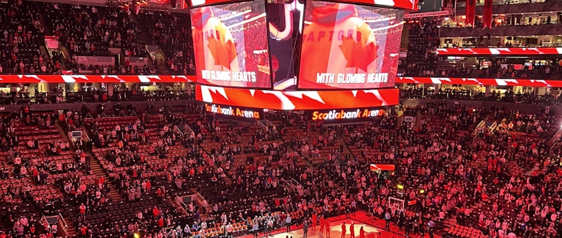 An indoor sports arena filled with spectators and a basketball court illuminated by red lighting. The scoreboards and video screens display promotional content, with numerous banners and championship flags hanging from the ceiling. Players and officials are gathered on the court, possibly in preparation for a game or event.
