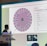 A person stands in front of a large screen displaying a colorful digital wheel with names. The screen shows a web interface with options for inputs, featuring a list of names with checkboxes on one side and a large, vibrant spinning wheel on the other. The environment appears to be indoors, possibly in a conference or presentation setting with a podium.