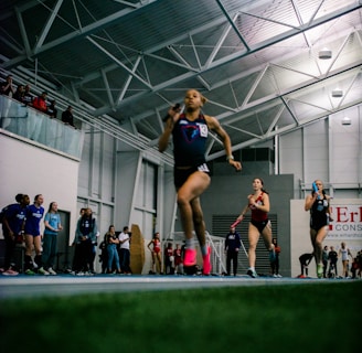 Athletes are competing in a relay race inside a large sports facility. The runners are moving swiftly along the track, with one runner in the foreground captured mid-stride. Spectators and other participants can be seen in the background, adding to the dynamic and energetic atmosphere of the scene.