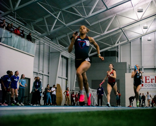 Athletes are competing in a relay race inside a large sports facility. The runners are moving swiftly along the track, with one runner in the foreground captured mid-stride. Spectators and other participants can be seen in the background, adding to the dynamic and energetic atmosphere of the scene.