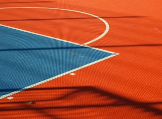 A vibrant sports court with sections of red and blue, featuring crisp white lines marking the boundaries. Shadows from a structure create geometric patterns across the surface, adding depth and interest to the scene.