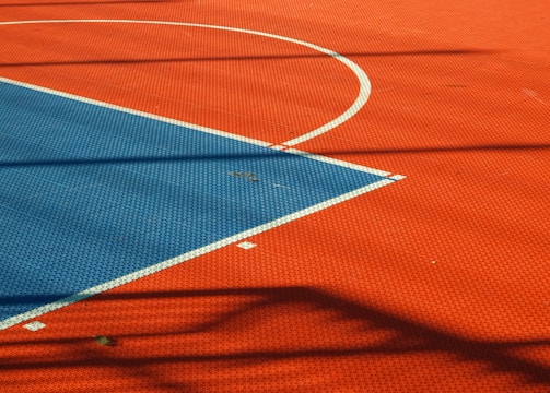 A vibrant sports court with sections of red and blue, featuring crisp white lines marking the boundaries. Shadows from a structure create geometric patterns across the surface, adding depth and interest to the scene.